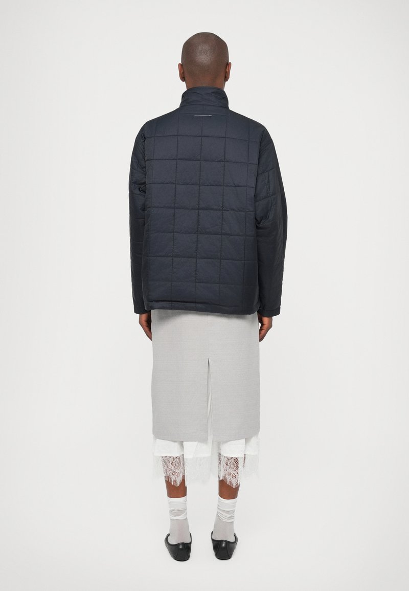 Person in einer schwarzen Steppjacke, einem hellgrauen Midirock mit einem Schlitz hinten, weißen Spitzenverzierungen darunter, weißen Socken und schwarzen Schuhen.