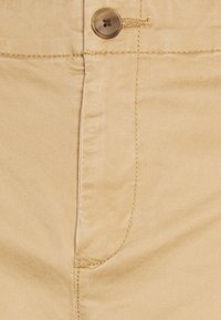 Chino in cotone beige con chiusura a bottone, cuciture in evidenza e texture liscia. Include dettagli delle tasche anteriori.