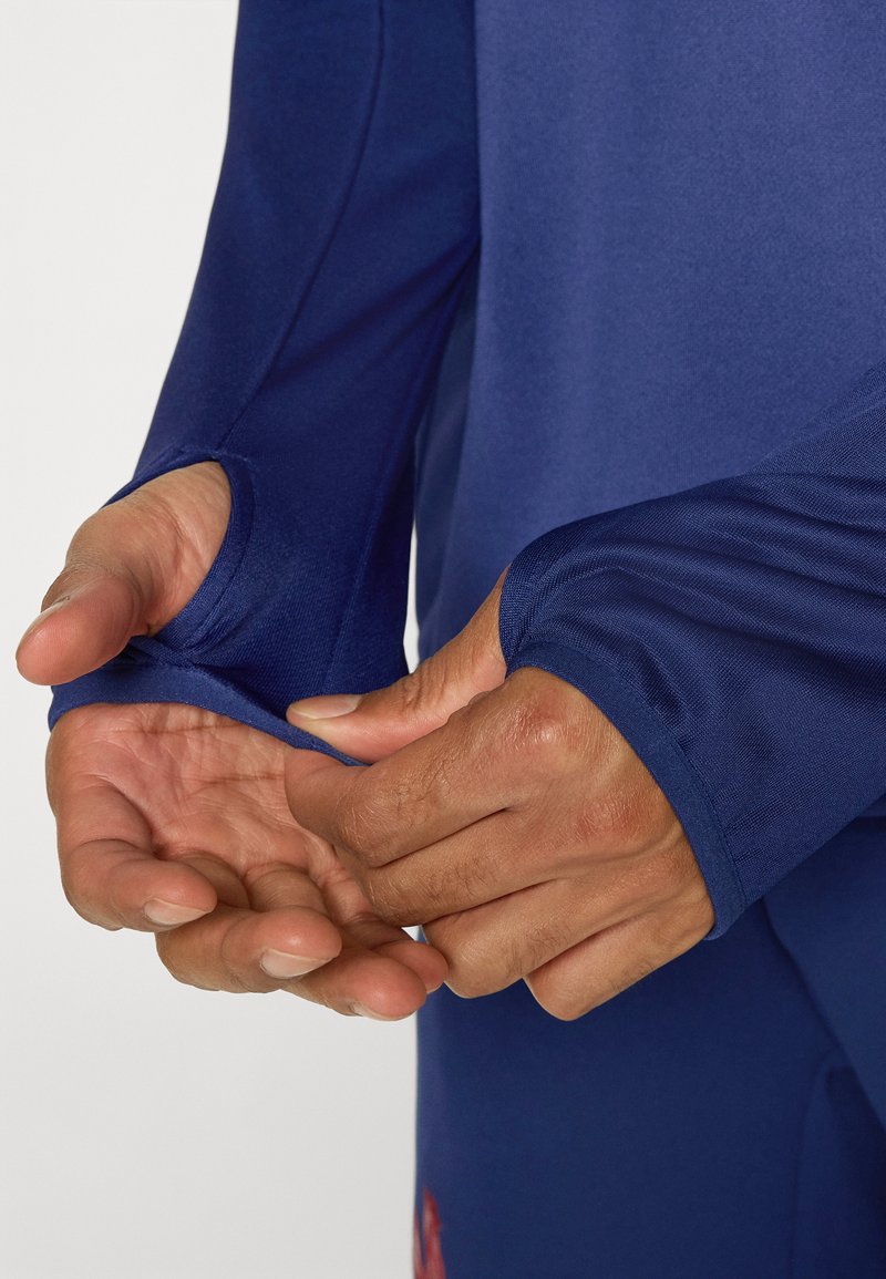 Haut de sport à manches longues en tissu bleu marine profond, doté de passe-pouces aux poignets pour un ajustement sûr et d'une finition lisse et texturée.
