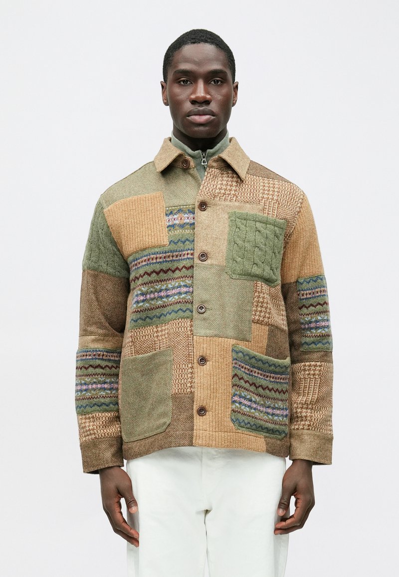 Casaco patchwork com texturas em verde, castanho e bege, apresentando padrões de tecidos mistos e fecho frontal de botões. Inclui gola com zíper e bolsos.