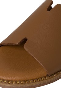 Hnědé kožené klouzací sandály s vyříznutým designem, texturovaným povrchem a zlatými kovovými nýty podél podrážky.