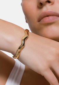 Glitrende gullfarget bølgete armbånd med en glatt, shiny finish. Har et minimalistisk design som forsiktig omfavner håndleddet.