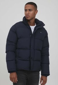 Marineblå jakke med dun og glidelås og trykknapper, høy krage og quiltet design. Stoffet ser glatt og isolert ut, med en logo-detalj.