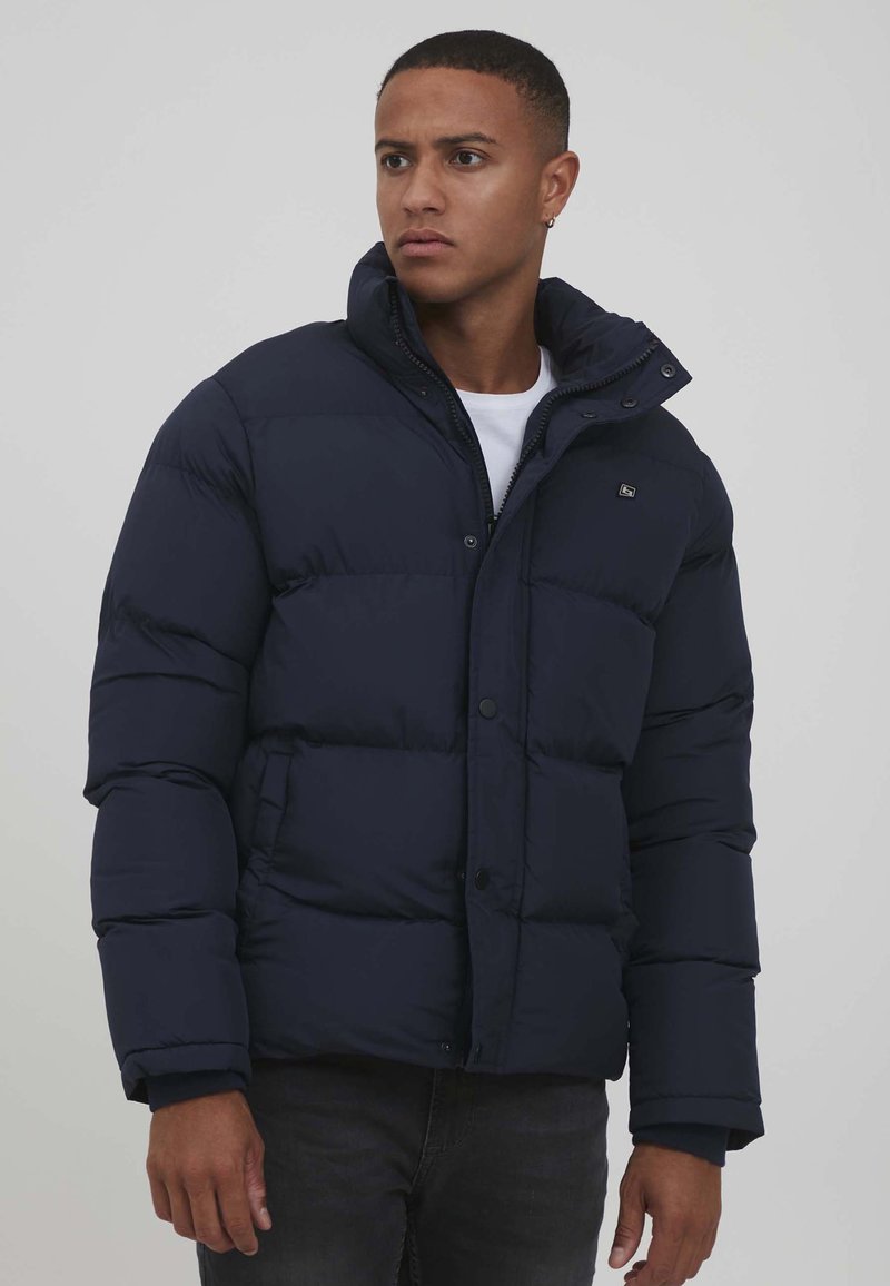 Marineblå jakke med dun og glidelås og trykknapper, høy krage og quiltet design. Stoffet ser glatt og isolert ut, med en logo-detalj.