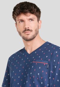 Camiseta de algodón azul marino con cuello en V, que presenta un patrón de anclas blancas, rojas y azules. Incluye un bolsillo con un borde en contraste.