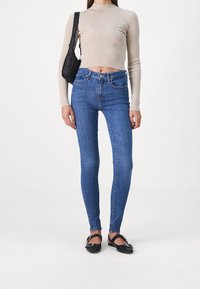 Mujer vestida con un top corto de manga larga beige, jeans ajustados de tiro alto azules, zapatos planos negros con hebilla y un bolso de hombro con correa de Tommy Jeans.