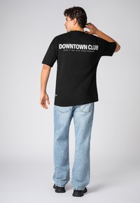 Schwarzes Baumwoll-T-Shirt mit weißem Text "DOWNTOWN CLUB" und kleinerem Text auf der Rückseite, kombiniert mit hellblauen Denim-Jeans und schwarzen Schuhen.