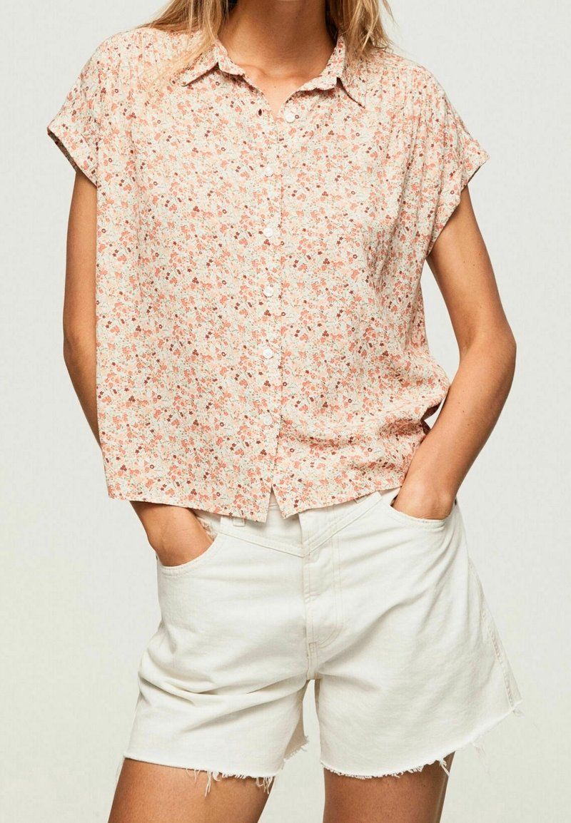 Femme portant un chemisier fleuri à manches courtes et boutonné, rentré dans un short en denim blanc taille haute avec ourlets effilochés, les mains dans les poches.