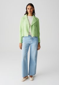 Giacca corta verde chiaro con texture, dotata di doppi bottoni e ampi rever, abbinata a jeans a gamba larga azzurri e scarpe beige.