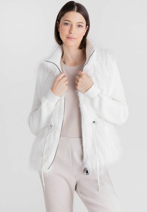 Vrouw met steil donker haar draagt een witte, pluizige jas met rits over beige geribbelde broek en top tegen een effen lichte achtergrond.