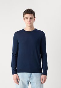 Sötétkék hosszú ujjú pulóver, kerek nyakkivágással, puha anyagú, testhezálló kialakítás. Világoskék farmernadrággal viselve, minimális logóval.