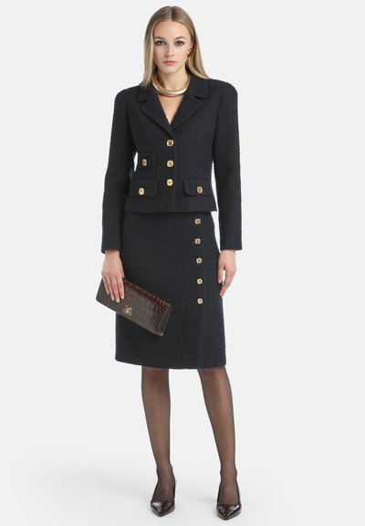 Woman wearing a black textured skirt suit with gold buttons, holding a dark brown clutch, and wearing black sheer tights and pointed heels.