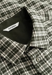 Col et bouton d’une chemise à carreaux verts et blancs avec une étiquette de marque blanche GABBA visible à l’intérieur du col.