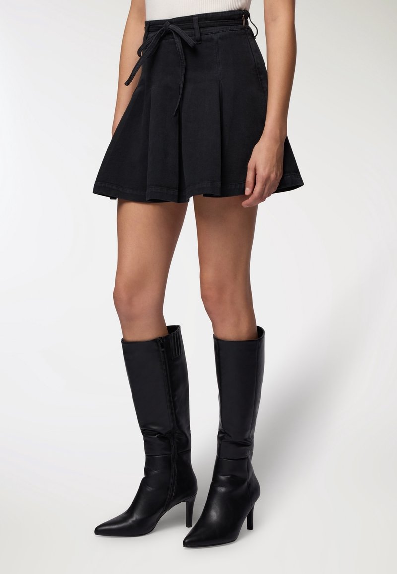 Femme portant une mini-jupe noire plissée avec une ceinture nouée à la taille et des bottes noires à talons montant jusqu'au genou sur un fond uni.