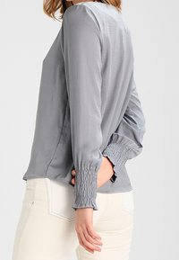 Blusa grigia a maniche lunghe con una texture liscia, arricciature sui polsini e una vestibilità rilassata, abbinata a pantaloni chiari.