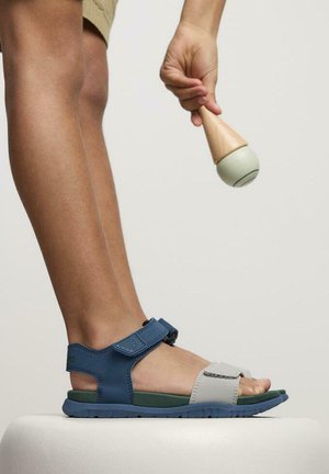 Sandalia azul y gris con cinta ajustable, suela texturizada y diseño de punta abierta. Usada en un pie bronceado, sosteniendo un juguete de madera.