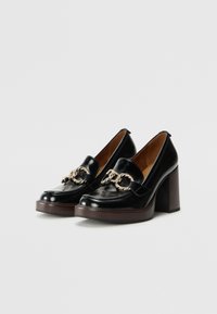 Mocassins à talons hauts en cuir noir avec un talon épais, dotés d'un détail décoratif en chaîne dorée et d'une finition brillante.