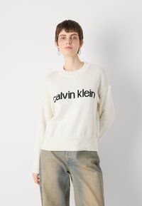 Hvit strikket genser med svart "calvin klein" logo over brystet, rund hals, og avslappet passform, kombinert med lysvasket baggy jeans.
