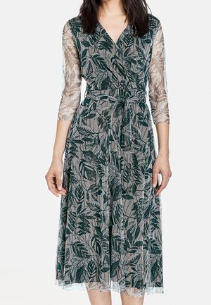 Mujer con un vestido cruzado hasta la rodilla con estampados de hojas verdes y mangas tres cuartos transparentes, ajustado a la cintura con un cinturón a juego.