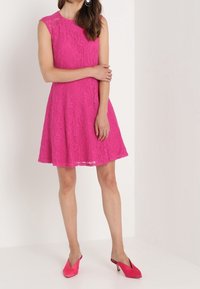 Ljust rosa spetsklänning med utsvängd kjol och korta ärmar, med ett blommönster. Kombinerad med matchande högklackade mules.