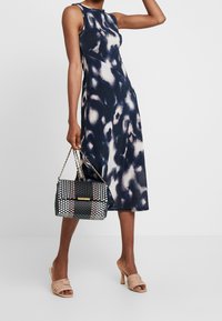 Abito senza maniche blu navy con un leggero motivo astratto, abbinato a una borsa a mano a scacchi bianco e nero e mule con tacco nudo.