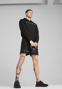 Hanorac negru și pantaloni scurți cu branding PUMA. Modelul ține o greutate kettlebell neagră, purtând pantofi sport negri. Pielea are tatuaje complexe.