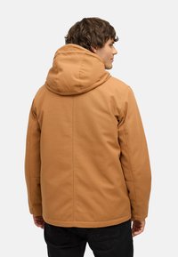 Brun hættejakke med en glat overflade, der har en skræddersyet pasform og minimal detaljering. Båret med mørke bukser, set fra ryggen.