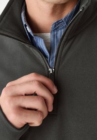 Tumši pelēka fleeces jaka ar rāvējslēdzēja aizdari. Iekšpusē redzama zilas un baltas svītraina apkakles krekla daļa. Tuva plaukstas tvertne ap rāvējslēdzēju.