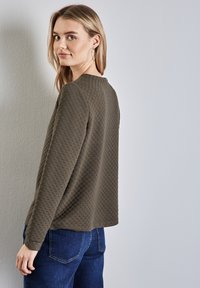 Olivengrön texturerad tröja med rund halsringning och långt ärm, som kombineras med mörkblå jeans. Suddig diamantmönster på tyget.