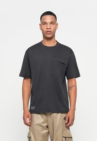 Ung mand iført en mørkegrå oversized T-shirt med en stor brystlomme og beige cargobukser, stående mod en ensfarvet hvid baggrund.