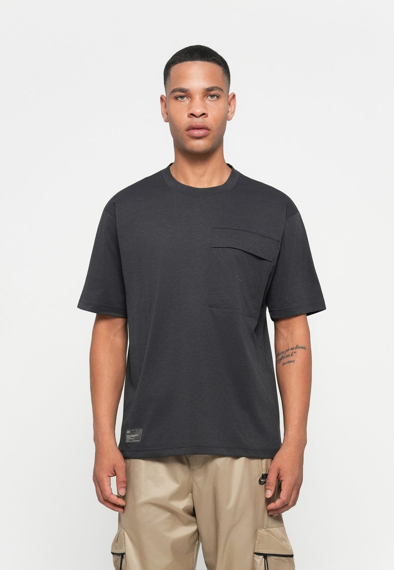 Ung mand iført en mørkegrå oversized T-shirt med en stor brystlomme og beige cargobukser, stående mod en ensfarvet hvid baggrund.
