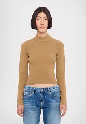 Ribbet, langermet høyhalset genser i beige, tettsittende design; brukt med blå denimjeans som viser en kort lengde.