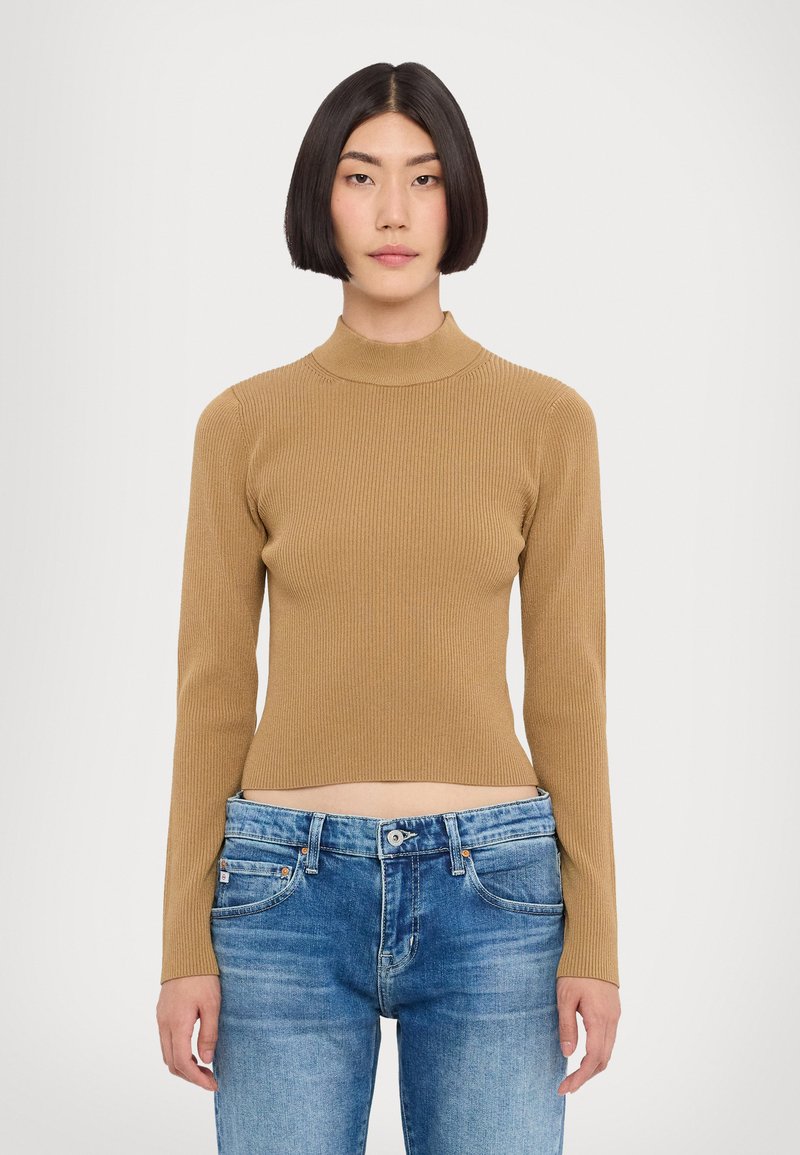Pull en maille côtelée, col roulé, à manches longues couleur beige, au design ajusté ; porté avec un jean en denim bleu, mettant en valeur une longueur courte.