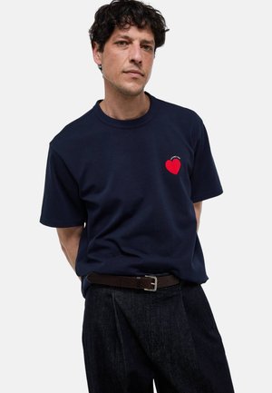 Homme portant un t-shirt bleu marine avec un cœur rouge et un texte, pantalon foncé et ceinture marron, debout devant un fond clair uni.