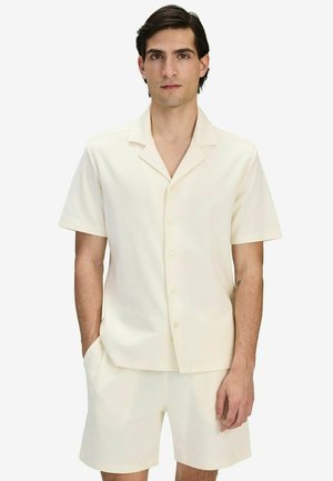 Mand iført kortærmet, cremefarvet skjorte med knapper og matchende shorts, stående med den ene hånd i lommen mod en hvid baggrund.