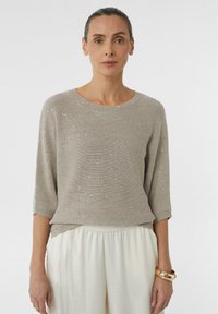 Beżowy sweter dzianinowy z okrągłym dekoltem, rękawami do łokcia i rozsianymi srebrnymi akcentami. Połączony z jasnobeżowymi spodniami.