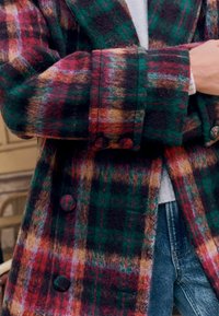 Villane ruuduline mantel, mis sisaldab punaseid, rohelisi ja pruune toone, suurte nööpide ja pehme tekstuuriga. Disain sisaldab struktureeritud kraed ja mansette.