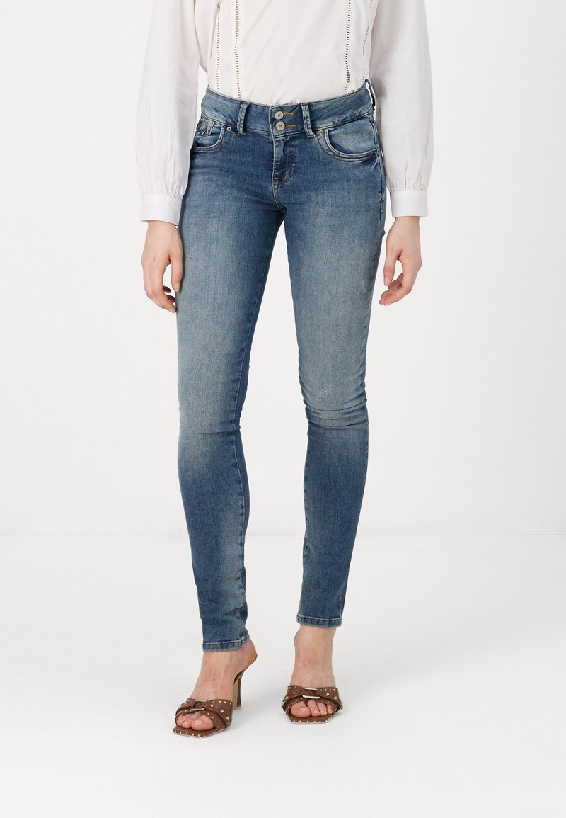 Jean skinny bleu en denim avec un délavage, deux poches avant et une taille haute, associés à des sandales à talons marron avec un détail de boucle.