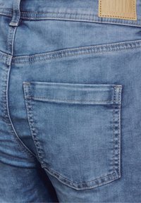 Blå denimjeans med en bakficka med subtila sömnadsdetaljer och en texturerad yta. En tan läderlogotyp är synlig.