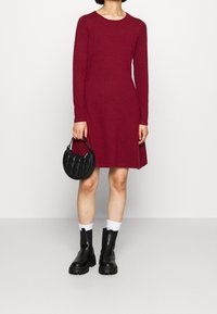 Maroon pitkähihainen mekko, jossa on pyöreä kaula-aukko ja liehuva helma, yhdistettynä mustaan tikattuun käsilaukkuun ja suuriin mustiin saapikkaisiin valkoisten sukkien kanssa.