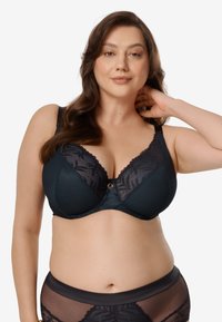 Kobieta o długich brązowych włosach, ubrana w ciemnoniebieski koronkowy biustonosz i dopasowane prześwitujące majtki, pozująca z jedną ręką za szyją na białym tle.