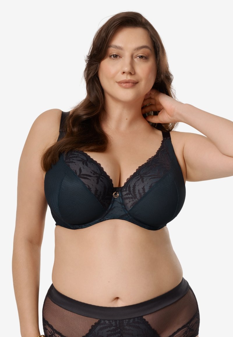 Kobieta o długich brązowych włosach, ubrana w ciemnoniebieski koronkowy biustonosz i dopasowane prześwitujące majtki, pozująca z jedną ręką za szyją na białym tle.