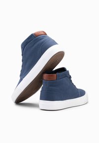 Zapatillas altas de lona azul oscuro con suelas de goma blanca y detalles en cuero marrón. Presentan cordones planos y paneles laterales texturizados.