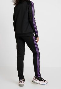 Svart sportig träningsoverall med lila ränder, med en avslappnad passform, ribbade muddar och matchande svarta sneakers med rosa inslag.