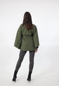 Olijfgroene jas in cape-stijl met wijde mouwen, een getailleerde taille met riem en een lichte textuur, gecombineerd met nauwsluitende grijze broek en zwarte enkellaarzen.