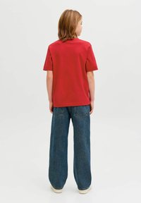 Vállig érő hajú gyermek piros rövid ujjú pólóban és bőszárú kék farmernadrágban, háttal áll egy sima háttér előtt.