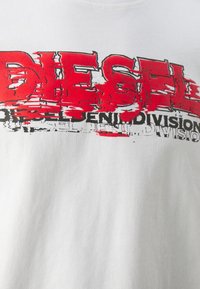 Bílý bavlněný tričko s výrazným, opotřebovaným červeným a černým potiskem "DIESEL". Kulatý výstřih, krátké rukávy a měkká textura.