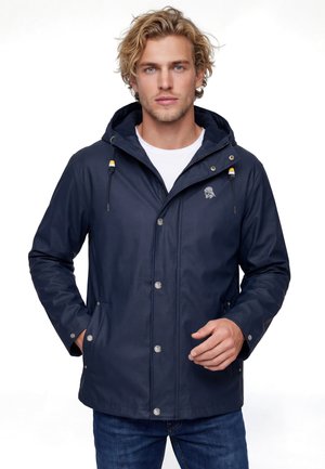 Giovane uomo che indossa una giacca blu navy con cappuccio sopra una maglietta bianca e jeans blu, in piedi con le mani in tasca contro uno sfondo bianco.