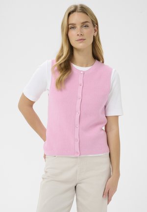 Woman with long blonde hair wearing a pink buttoned knit vest over a white short-sleeve shirt and light beige pants stands against white background.