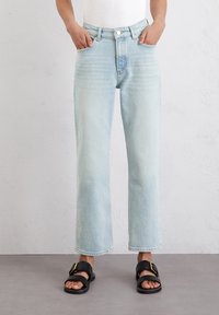 Persona con jeans rectos de color azul claro y sandalias negras con hebillas doradas, de pie contra una pared blanca texturizada.